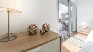 A modern bedroom with a wooden bedside table, a lamp, decorative eggs, a sliding glass door to a balcony, and a small wooden nightstand with a lamp. - Marina's Flat Limassol (Limassol)