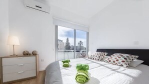 A modern bedroom with a large bed, a bedside table with a lamp, and a view of buildings through a large window. - Marina's Flat Limassol (Limassol)