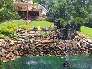 A pond with a waterfall, surrounded by rocks and greenery, with outdoor seating and a building in the background. - Private country hillside setting surrounded by trees, birds, wildlife pond/falls (Nashville)