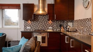 A compact kitchen with dark wooden cabinets, a tiled backsplash, a built-in oven, and a window with blinds. - Fabulous Apartment near Edinburgh Center (Edinburgh)