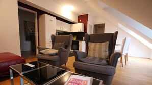 Un salon moderne avec deux fauteuils gris, une table basse en verre et un pouf rouge. On aperçoit une cuisine à l’arrière-plan, avec des meubles blancs et un plan de travail noir.