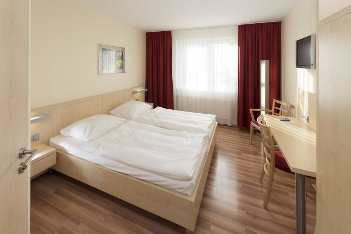 Ett hotellrum med en dubbelsäng, ett skrivbord, en stol, en TV och ett fönster med gardiner.