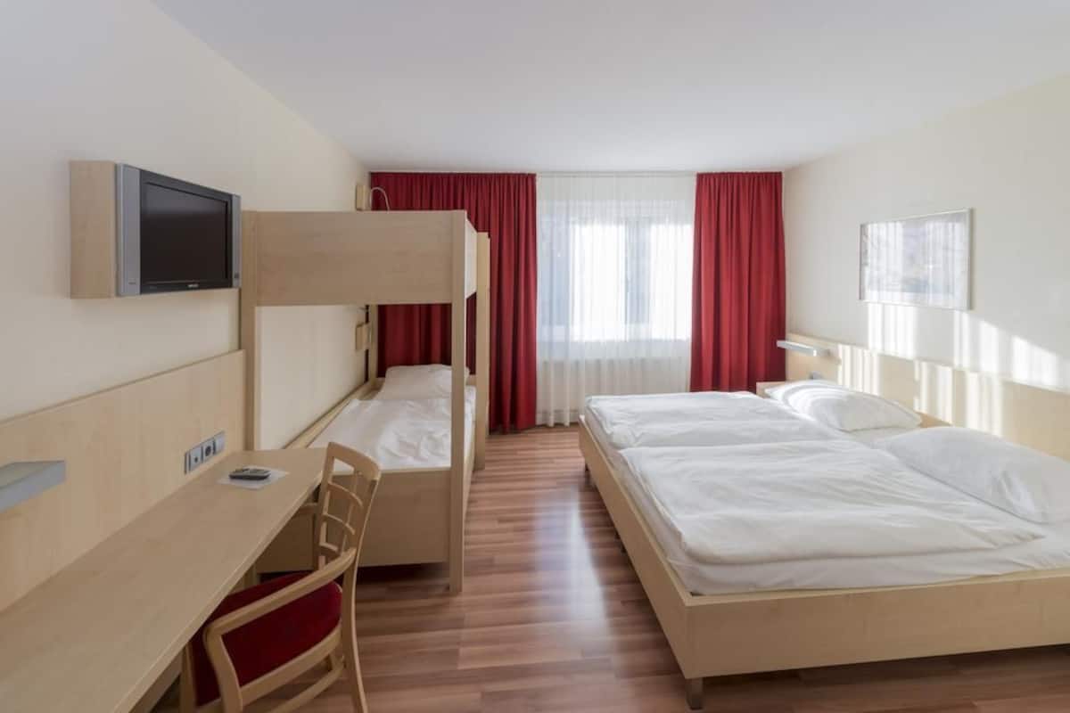 Ett hotellrum med en våningssäng, ett skrivbord med en stol, en tv och ett fönster med röda gardiner.