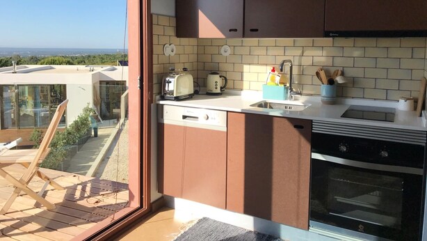 A modern kitchen with a built-in oven, a toaster, and a kettle. There is a sliding glass door leading to a balcony with a view of the landscape.