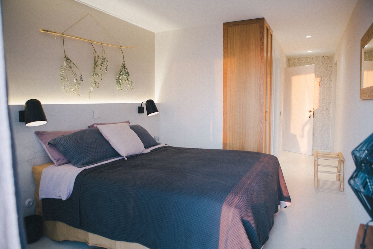 Ein Schlafzimmer mit einem Bett, Nachttischlampen, einer hängenden Pflanze, einem Holzschrank und einem kleinen Holzschemel.