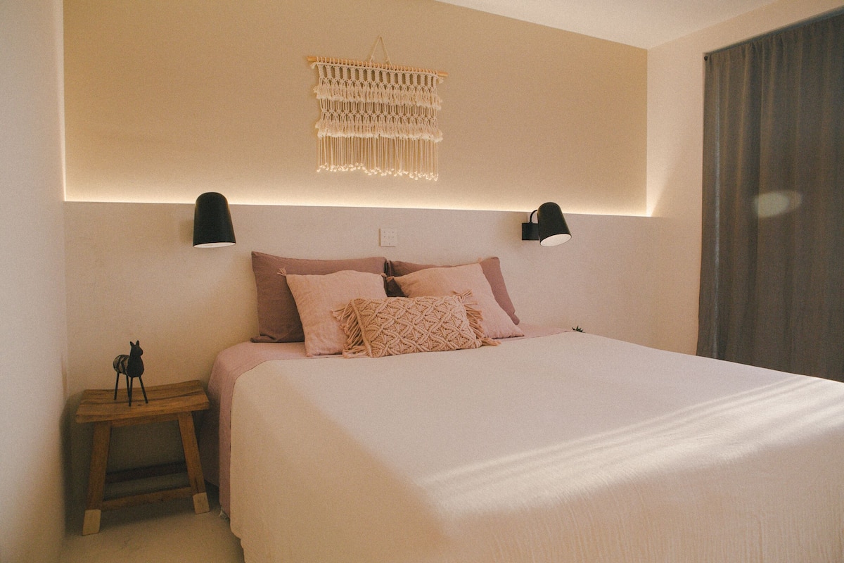 Ein Schlafzimmer mit einem Bett, Nachttischlampen, einem Wandbehang aus Makramee und einem kleinen Nachttisch mit einer Figur.