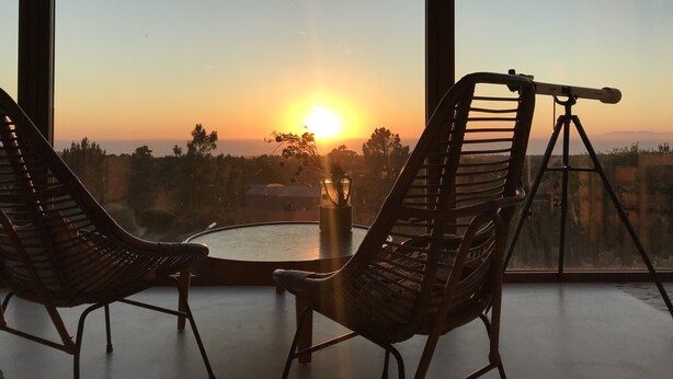 Una habitación con dos sillas de mimbre orientadas hacia una ventana grande con vistas a un atardecer.