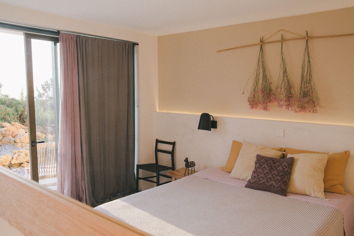 Ein Schlafzimmer mit einem Bett, einem Sessel, einem Fenster mit Vorhängen und einer Wandpflanze.