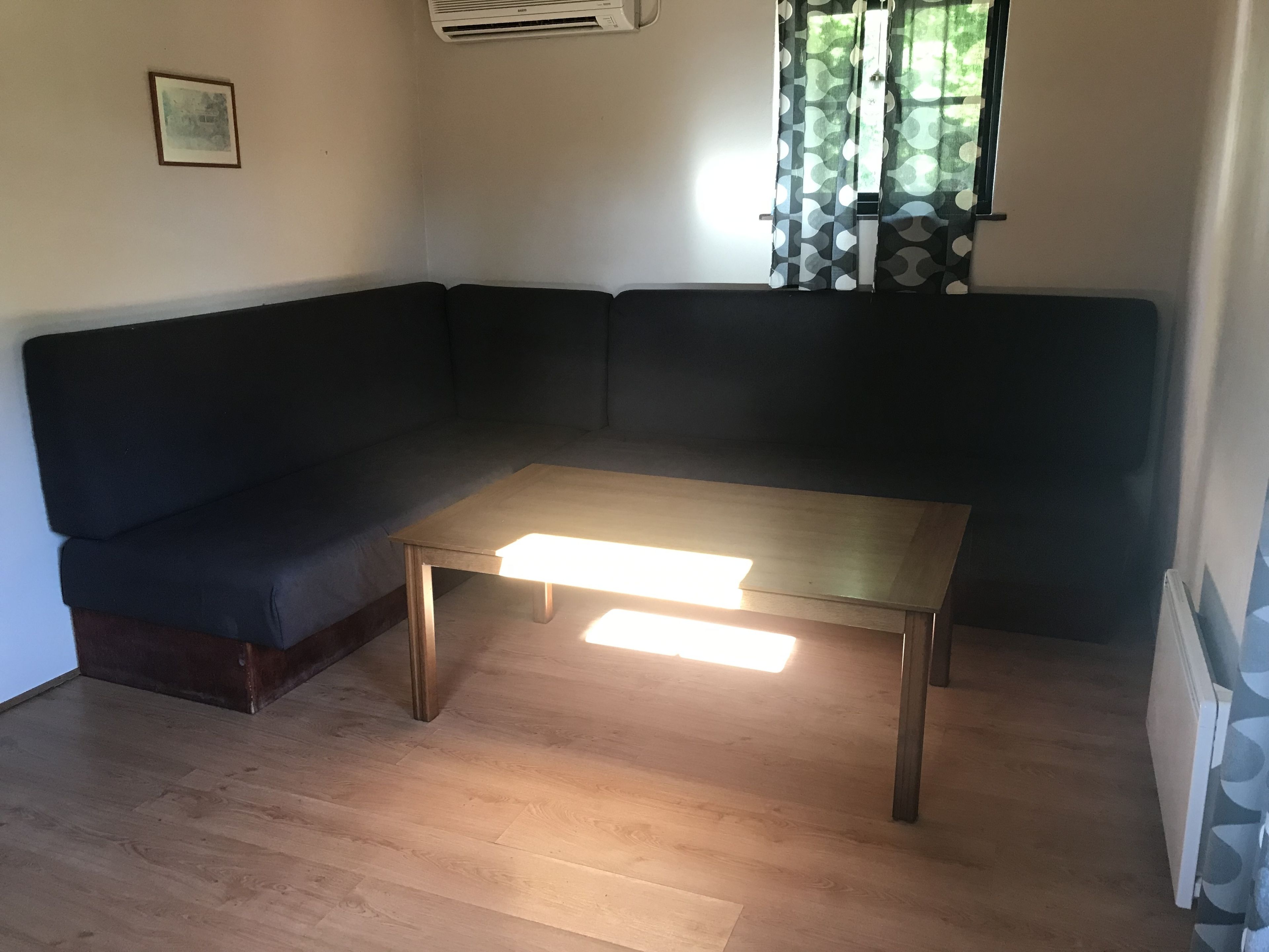 Une pièce d’angle avec un canapé de couleur sombre, une table en bois, une fenêtre ornée de rideaux à motifs et un tableau encadré accroché au mur.