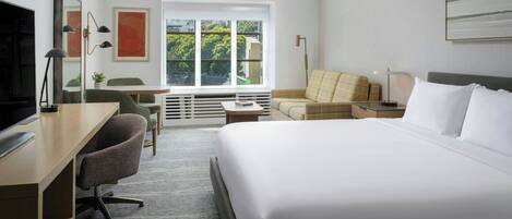 A hotel room with a bed, desk, chair, sofa, and a large window with a city view.