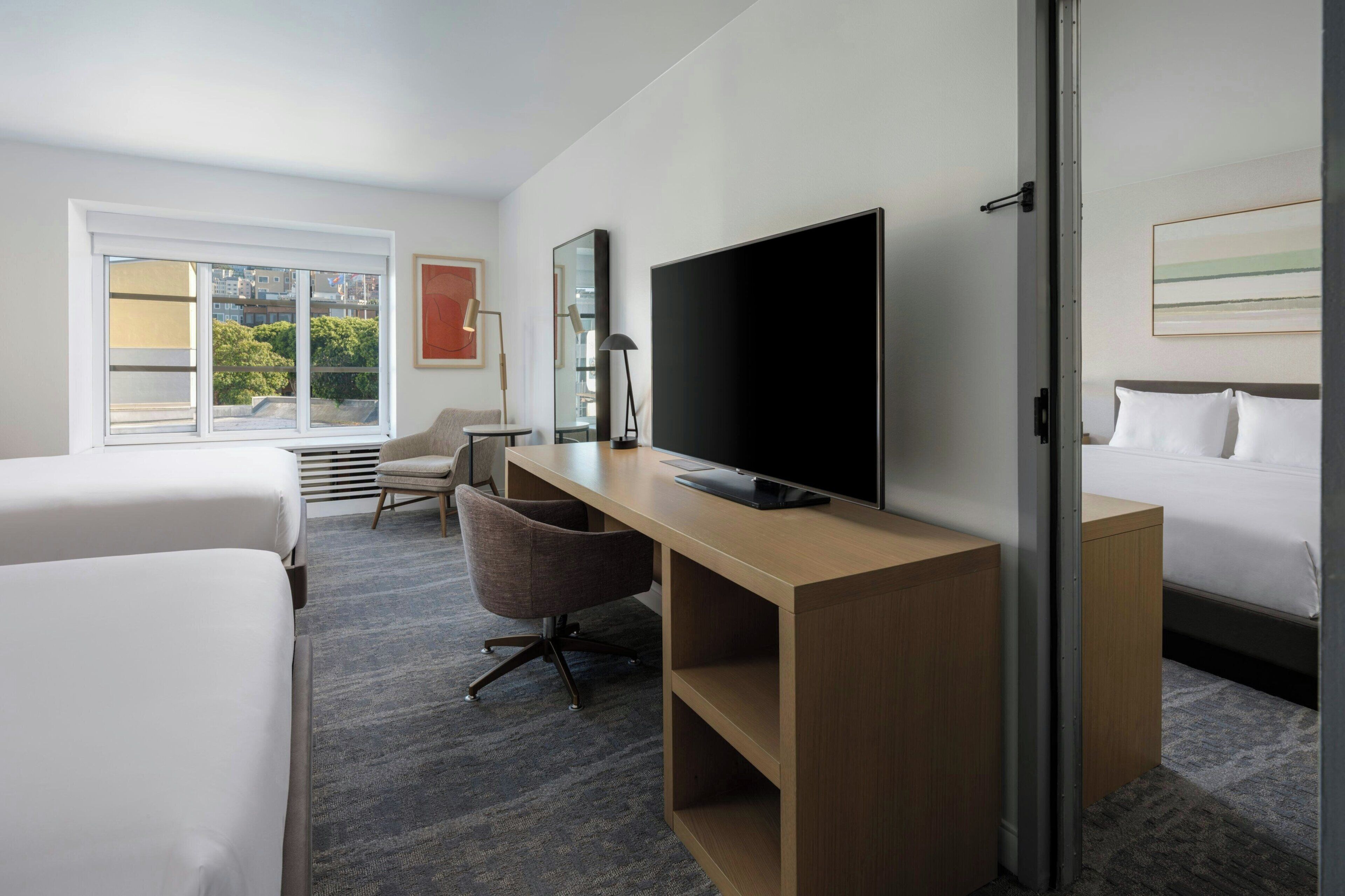 Habitación de hotel con cama, escritorio con televisor, silla y ventana con persianas.