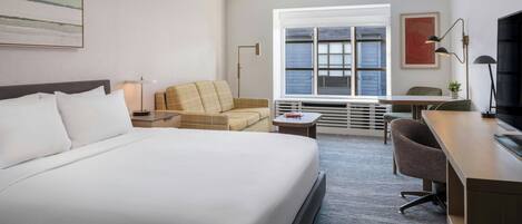 A hotel room with a large bed, a desk, a chair, a sofa, and a window with blinds.