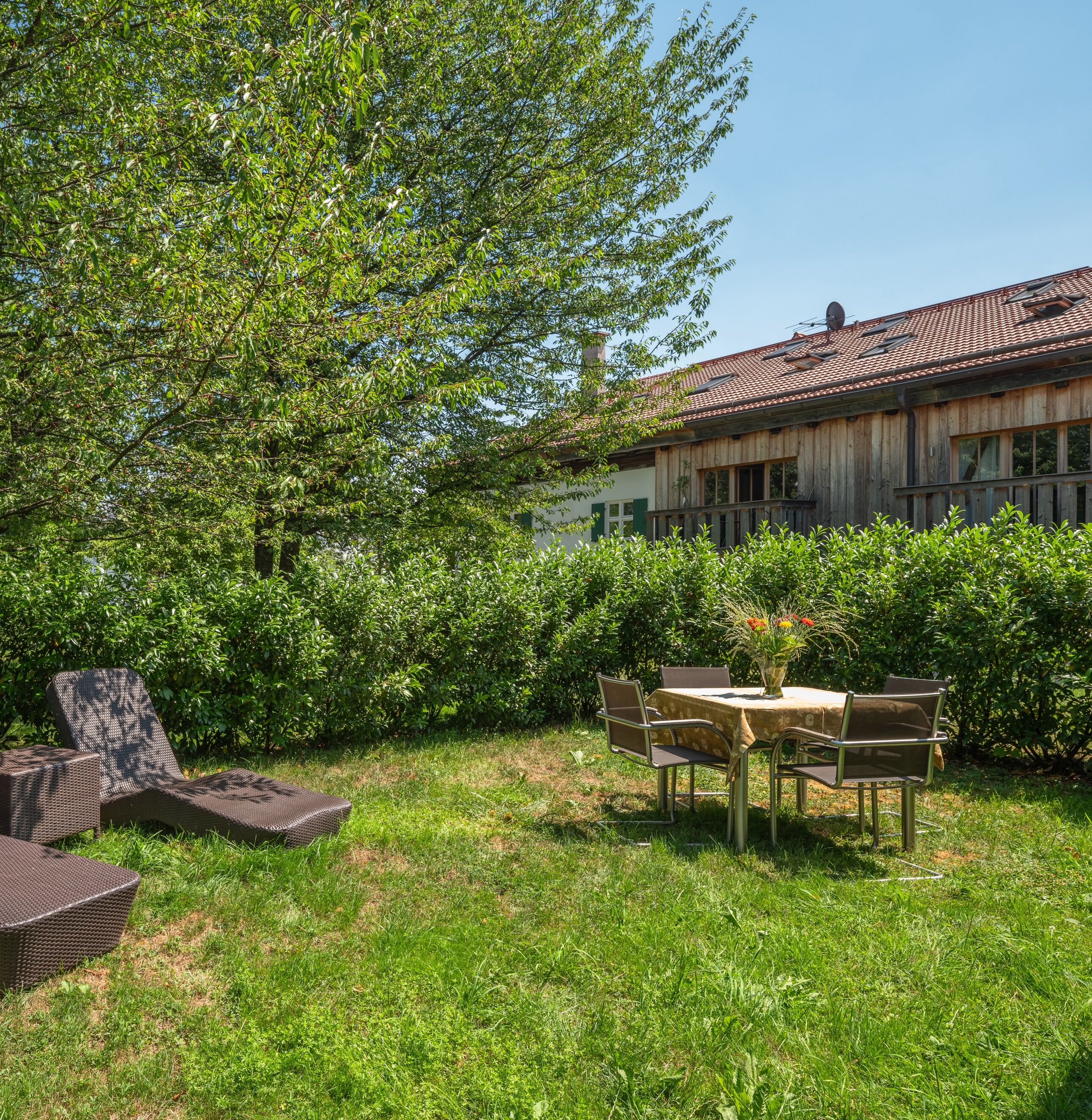 Um jardim com uma mesa e cadeiras, uma poltrona de vime e um edifício com telhado de telhas.