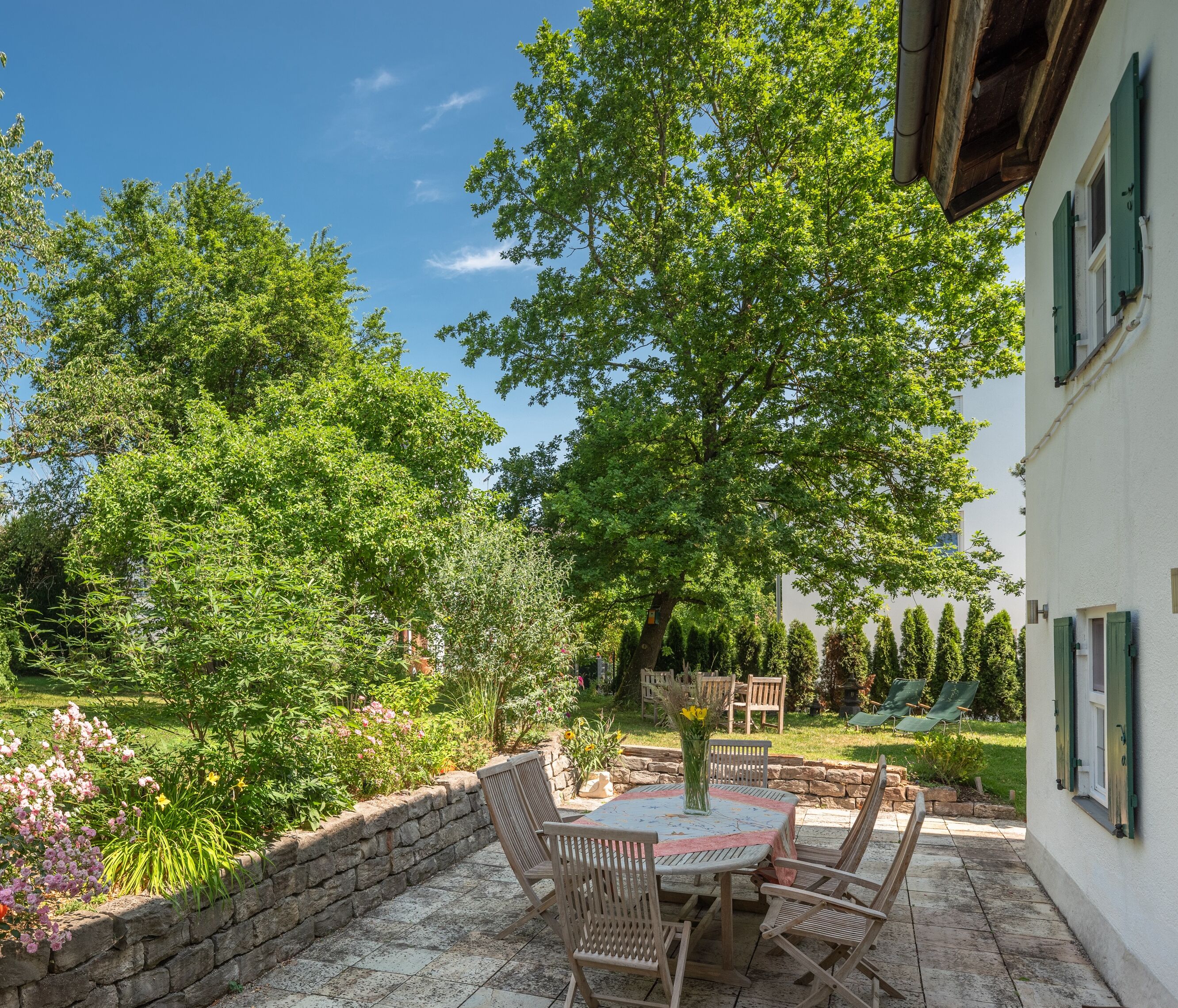A patio with a round table and chairs, surrounded by a stone wall and lush greenery.