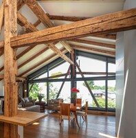 Um quarto espaçoso com teto alto, vigas de madeira, uma mesa de jantar, cadeiras, um sofá e grandes janelas com vista para o verde lá fora.