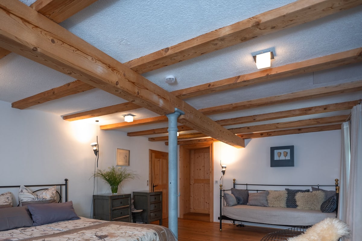 A bedroom with wooden beams, a bed, a sofa, a desk, and a plant.