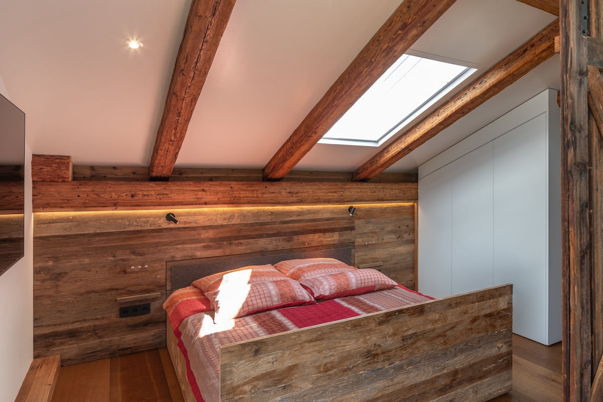 A bedroom with a sloped ceiling, wooden beams, a skylight, and a bed with patterned bedding.