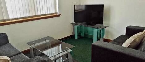 A living room with a flat-screen TV on a stand, a glass coffee table, and two grey sofas.