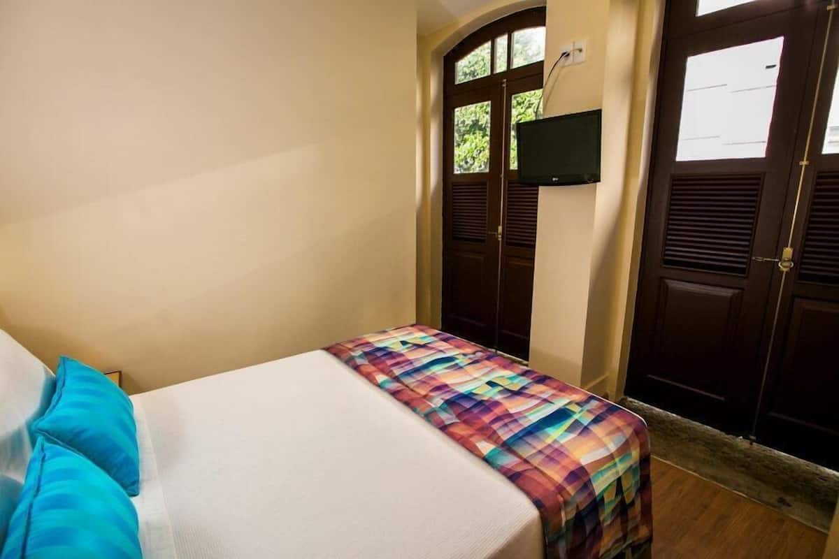 Una habitación pequeña con una cama individual, colcha escocesa, televisor montado en la pared y dos puertas con ventanas.