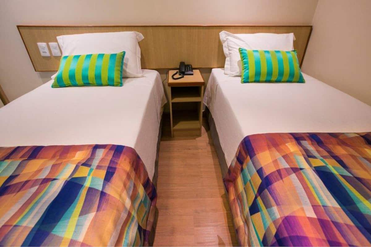Habitación de hotel con dos camas, cada una con una colcha de colores y un cojín verde a rayas. Entre las camas hay una mesita de noche de madera con un teléfono encima.