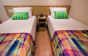 Habitación de hotel con dos camas, cada una con una colcha de colores y un cojín verde a rayas. Entre las camas hay una mesita de noche de madera con un teléfono encima.