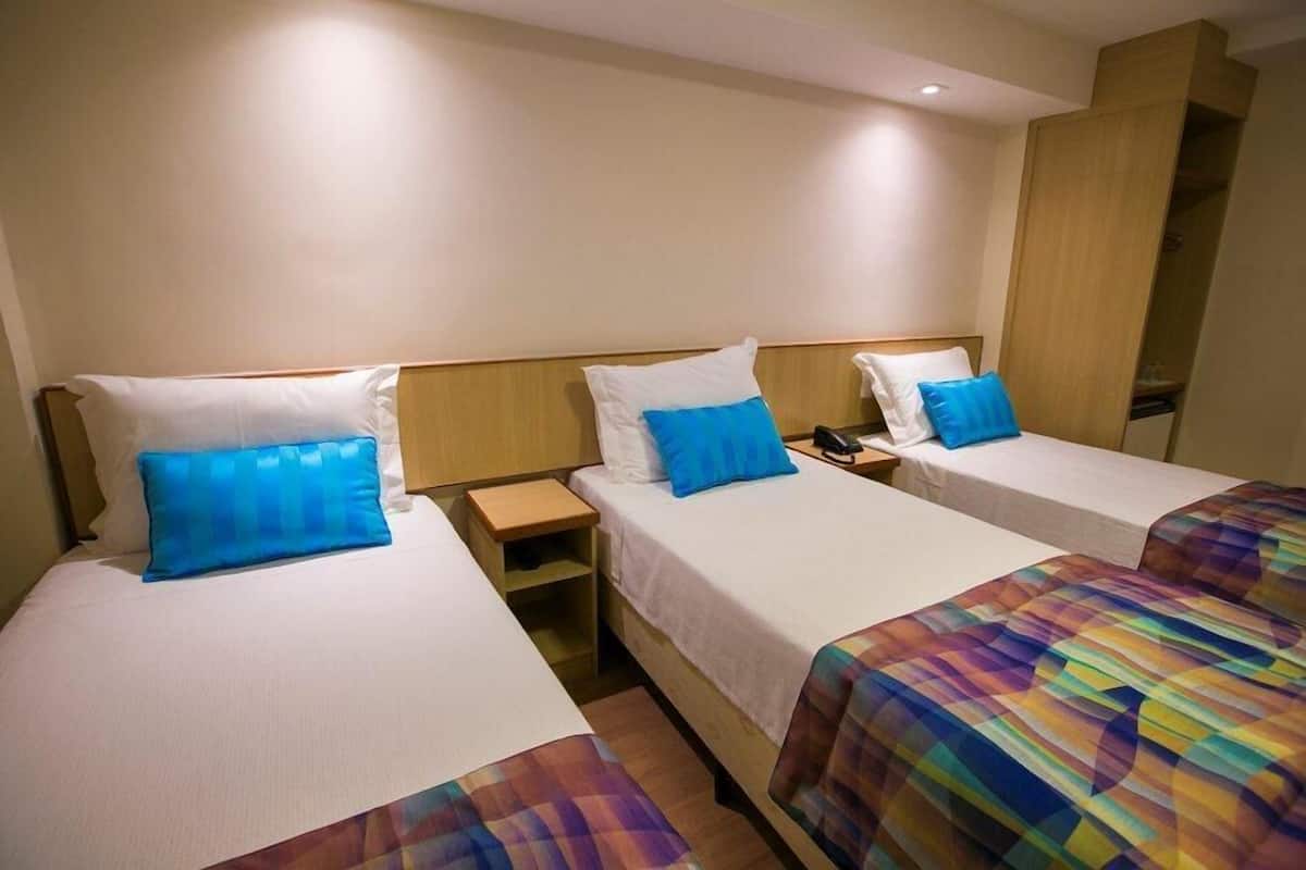 Habitación de hotel con tres camas individuales, cada una con colchón blanco y almohadas azules. Hay mesitas de noche de madera con teléfonos y lámparas.