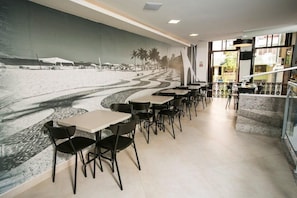 Un restaurante con un mural de una escena de playa, mesas y sillas dispuestas a lo largo de la pared, y una escalera que conduce a un nivel superior.