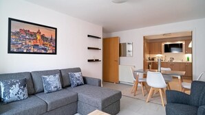 Ein modernes Wohnzimmer mit grauem Sofa, einem Esstisch mit Stühlen, einer Küchenzeile mit Schränken und einem Fernseher, sowie einem Stadtbild an der Wand.