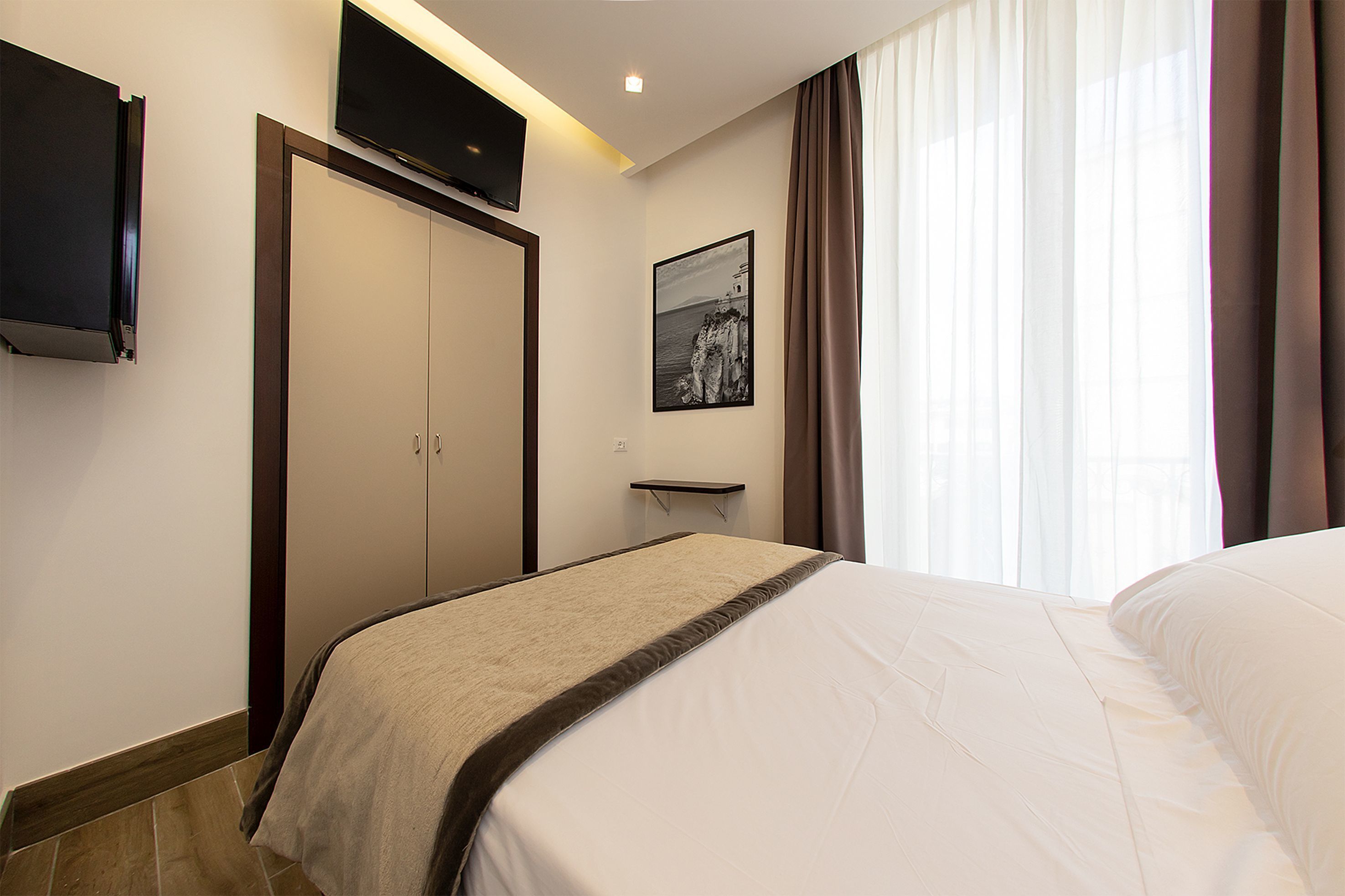 Une chambre d’hôtel avec un lit, un téléviseur à écran plat fixé au mur, un tableau encadré au mur et une fenêtre avec des rideaux.