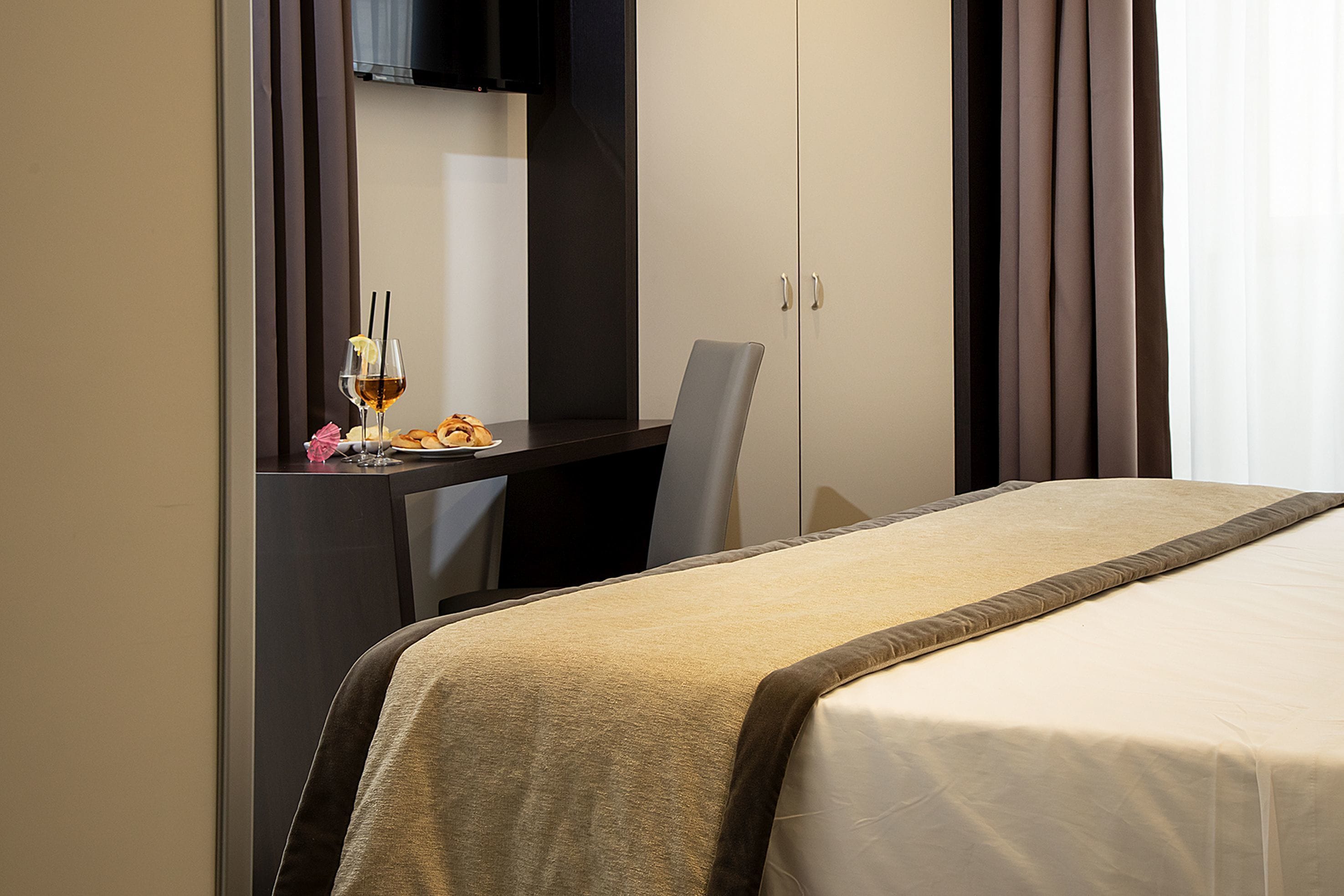A hotel room with a bed, a desk, and a chair. There is a glass of wine on the desk.