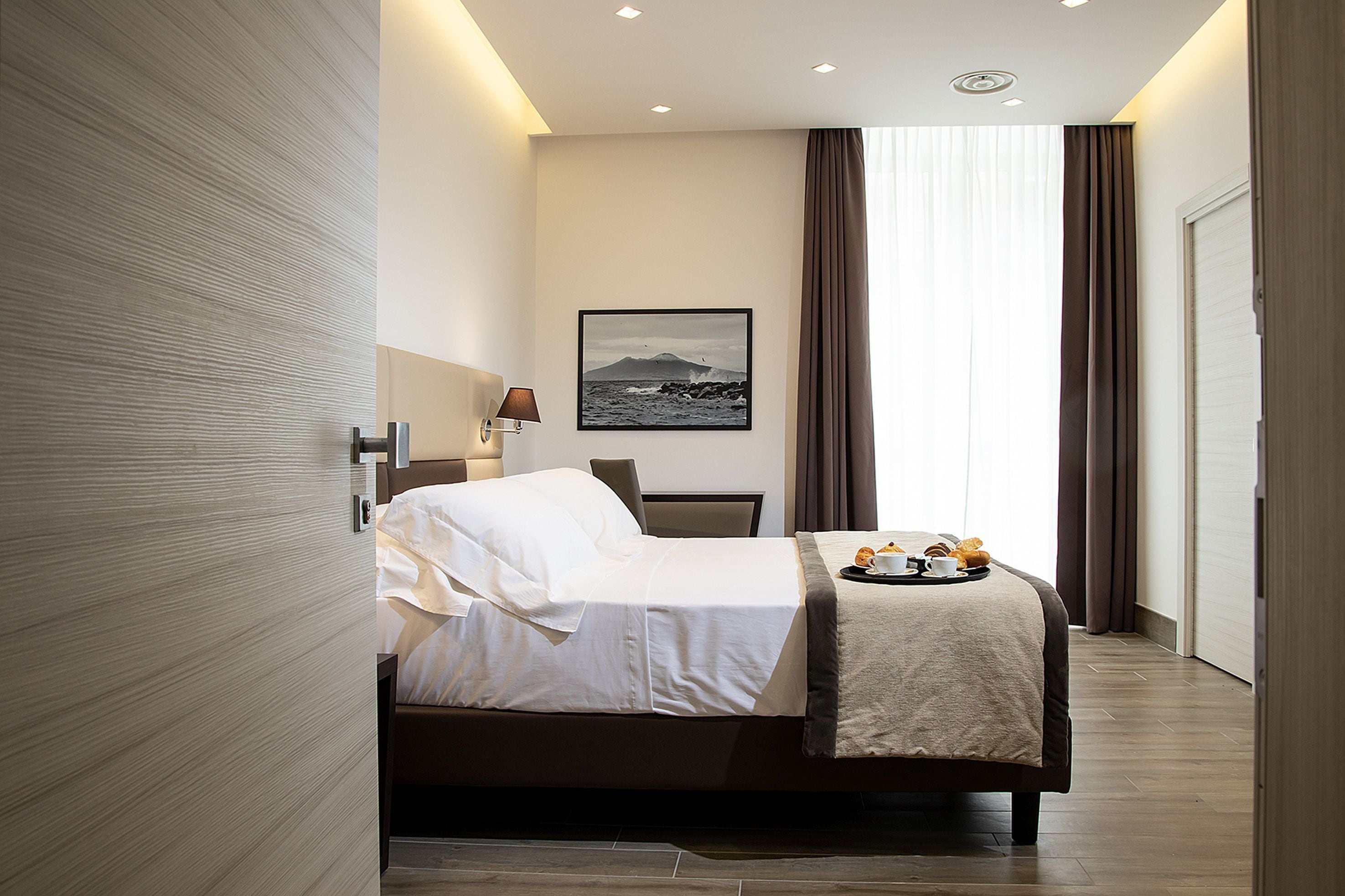 Una camera d'albergo moderna con un letto rifatto, un'area pranzo con un vassoio per la colazione, un quadro appeso alla parete e un'ampia finestra con tende.