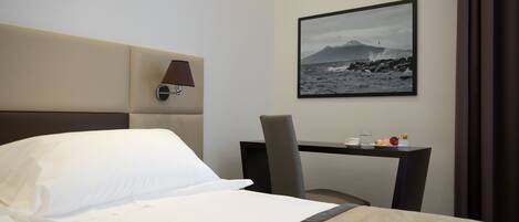 A hotel room with a bed, a desk, a chair, a lamp, and a framed picture of a coastal landscape.