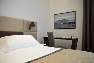 Una camera d'albergo con un letto, una scrivania, una sedia, una lampada e un quadro incorniciato con un paesaggio costiero.