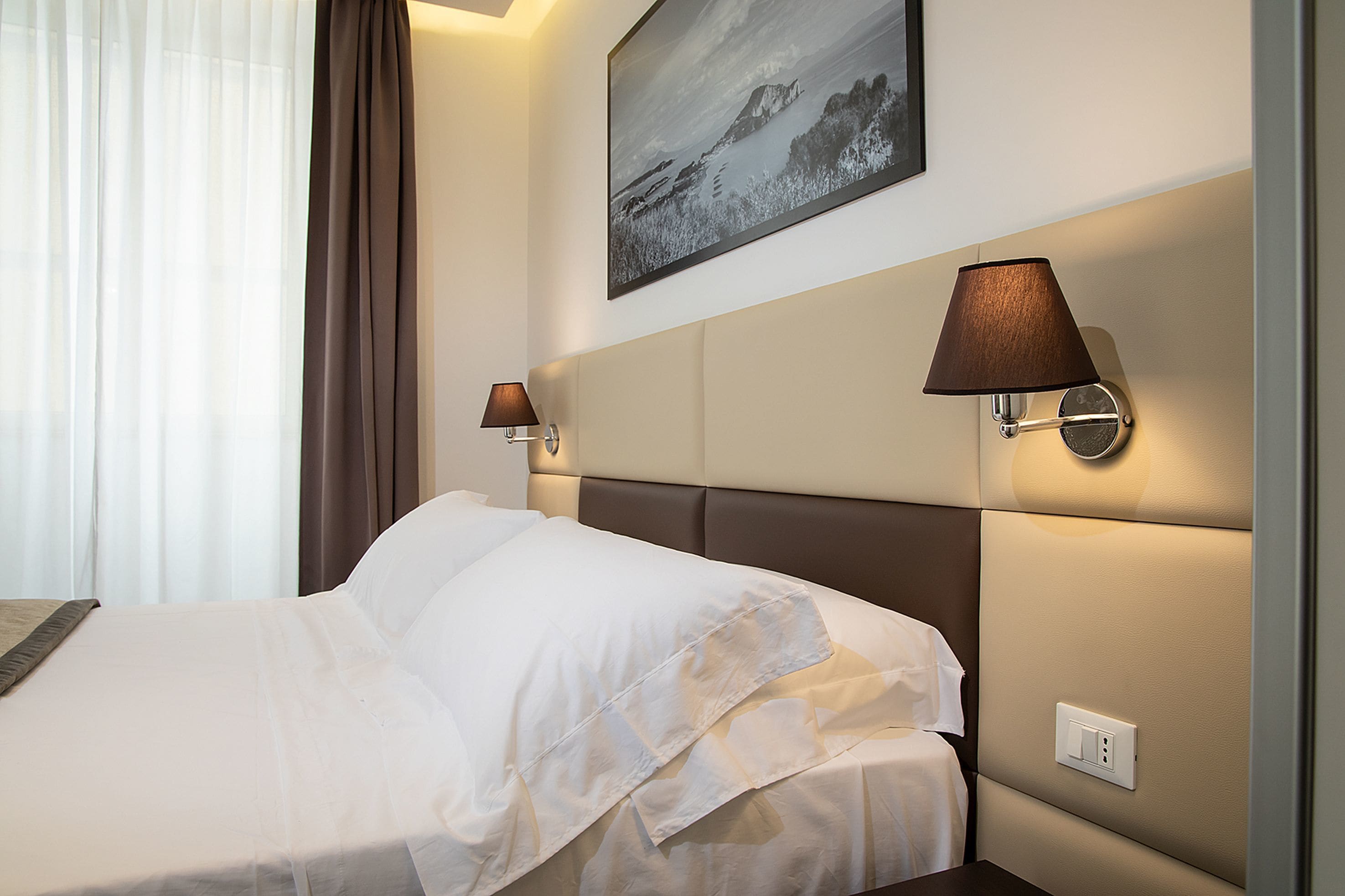 A hotel room with a large bed, two bedside lamps, a headboard, a painting on the wall, and a window with curtains.