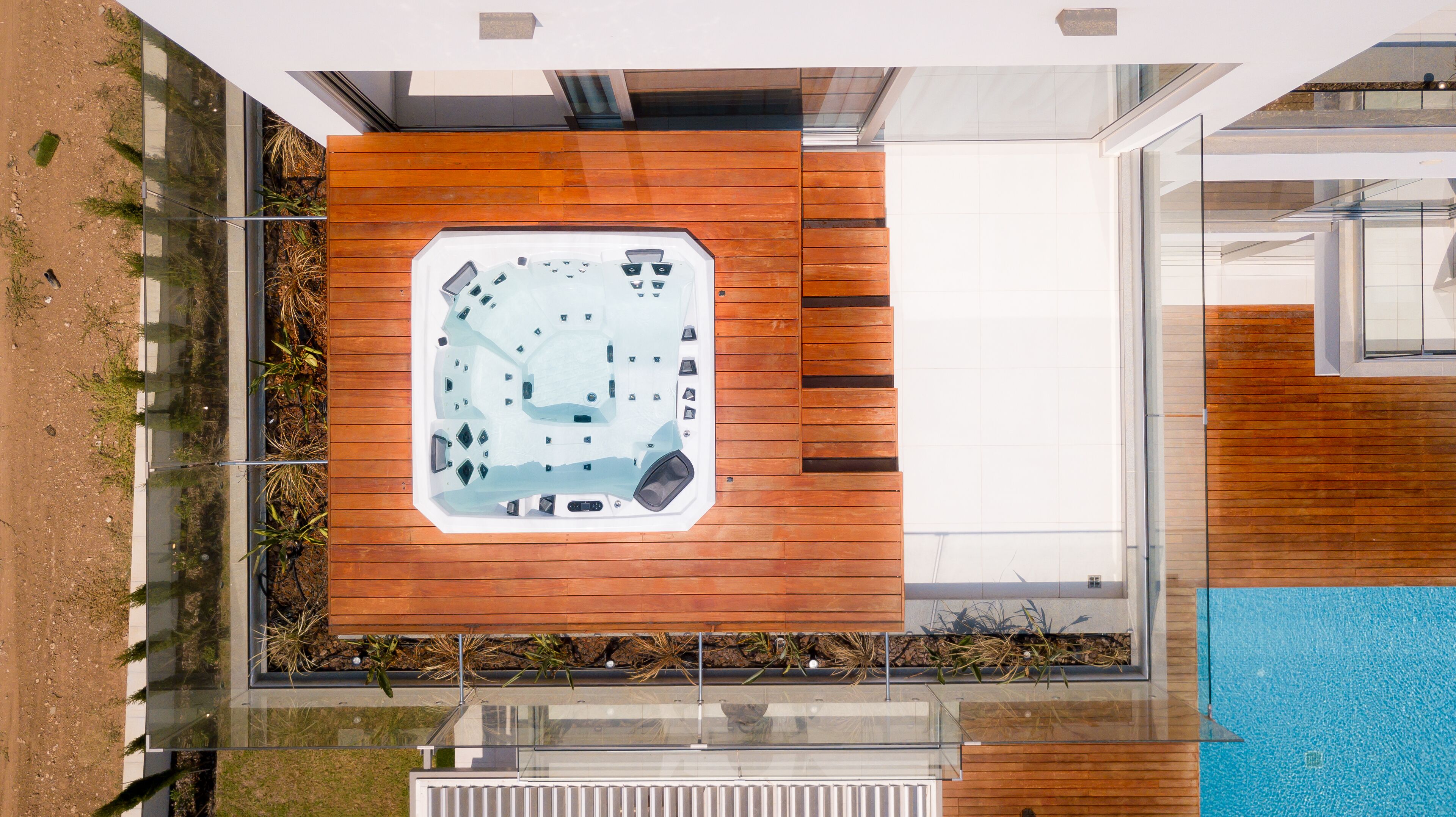 Un espace moderne aménagé à l’extérieur avec un jacuzzi, une terrasse en bois et des murs en verre, entouré d’une piscine et de verdure.