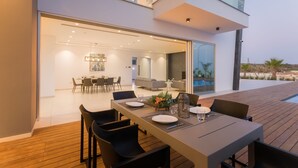 A modern outdoor dining area with a wooden deck, a long table set for six, and a pool visible in the background. - Cove Seaview Villas (Ayia Napa)
