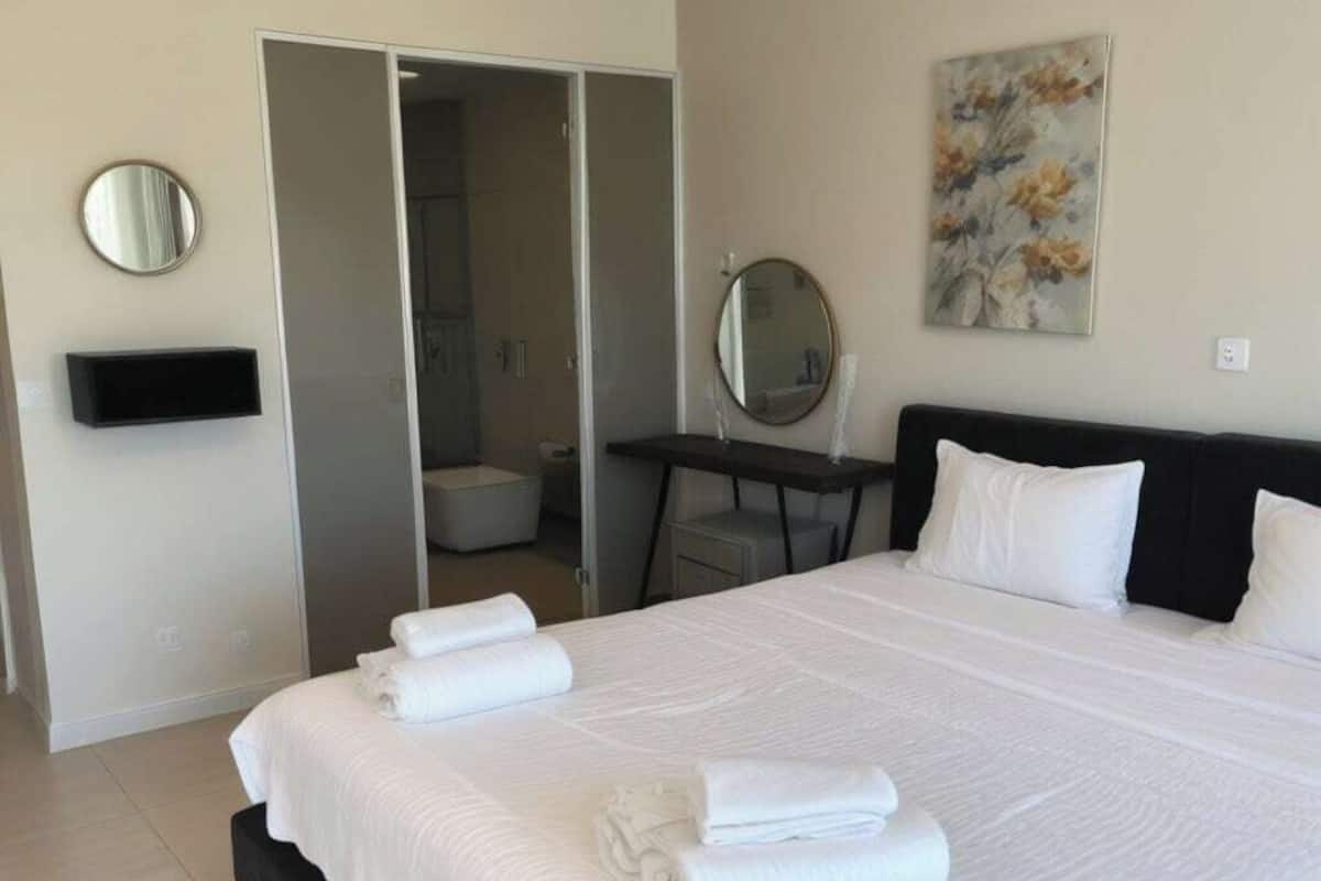 A modern hotel room with a large bed, a bedside table with a mirror, a small desk, and a bathroom visible through a sliding door.