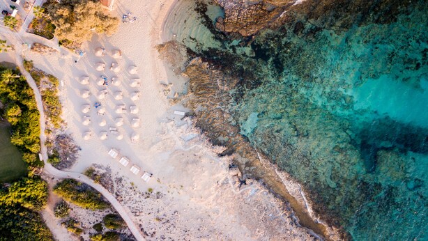 An aerial view of a beach with sun loungers and umbrellas arranged in rows, a clear path leading to the beach, and rocky areas with clear water. - Cove Seaview Villas (Ayia Napa)