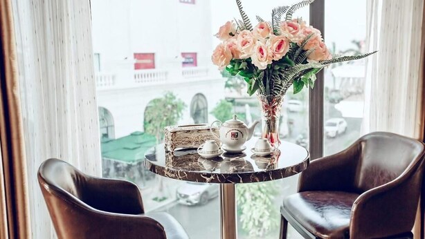 A corner table with a vase of flowers, a teapot, cups, and a box, set against a window with a view of buildings and cars. - The Oriental Ruby (Dong Hoi)