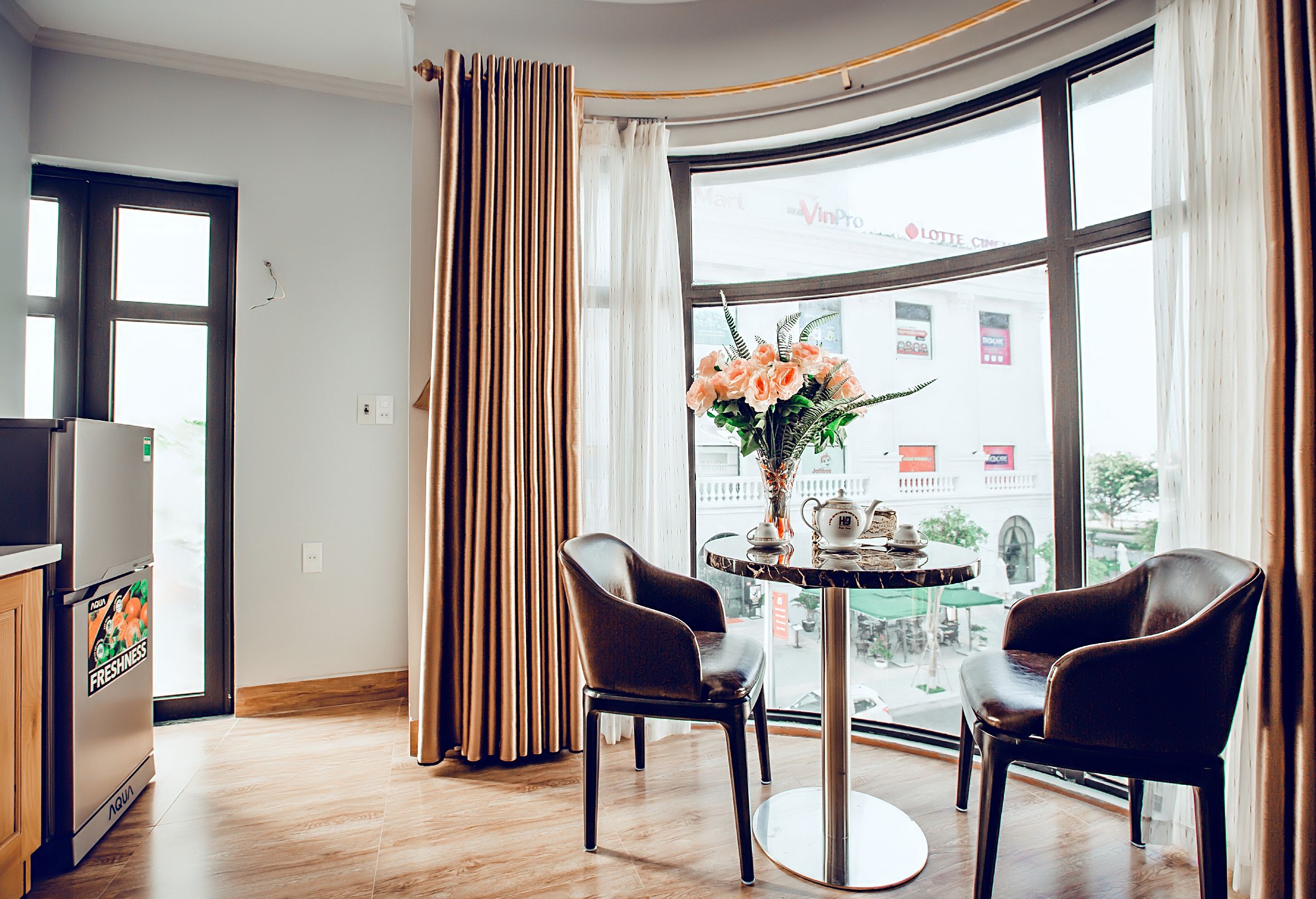 A modern hotel room with a large window, a dining table with chairs, and a refrigerator.
