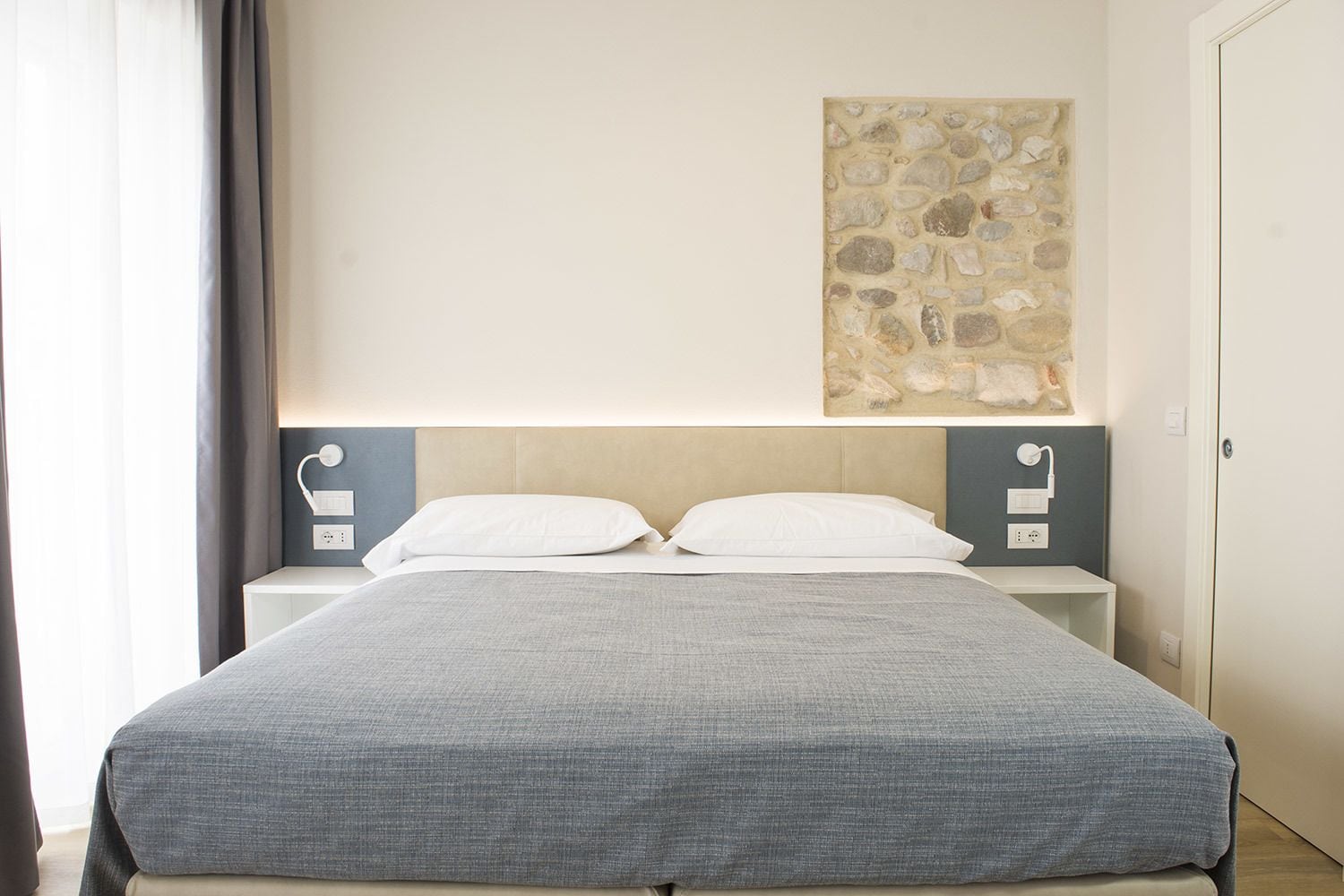 A neatly made bed with a grey bedspread, two white pillows, and a headboard with a stone wall art piece above it.