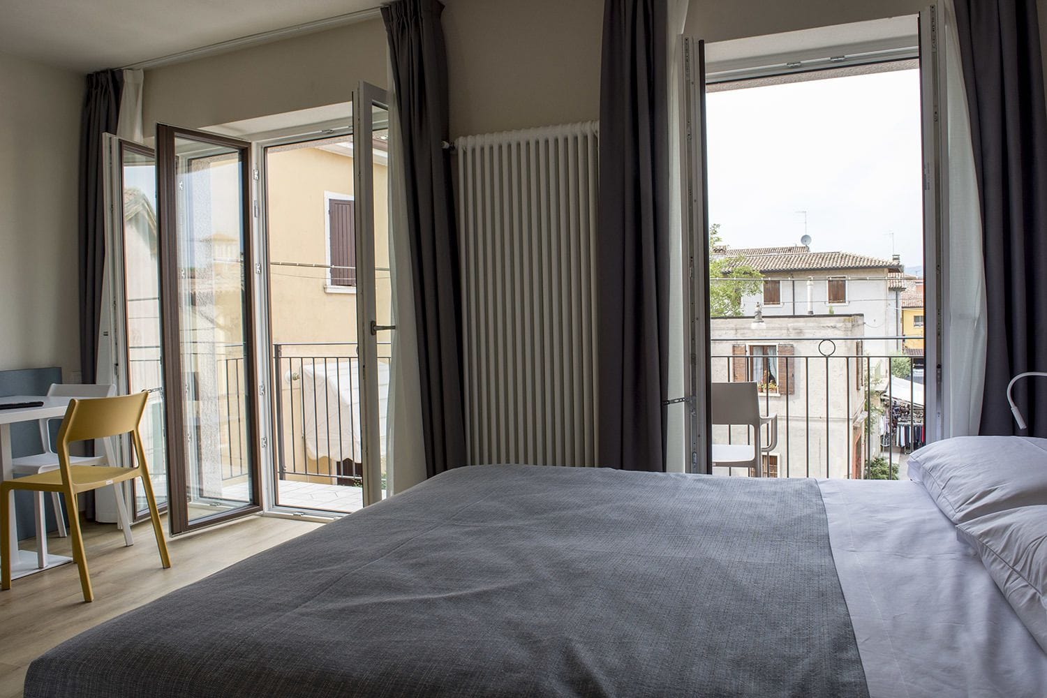 Une chambre d’hôtel avec un lit, un bureau et une chaise. Elle dispose d’un balcon donnant sur les bâtiments environnants.