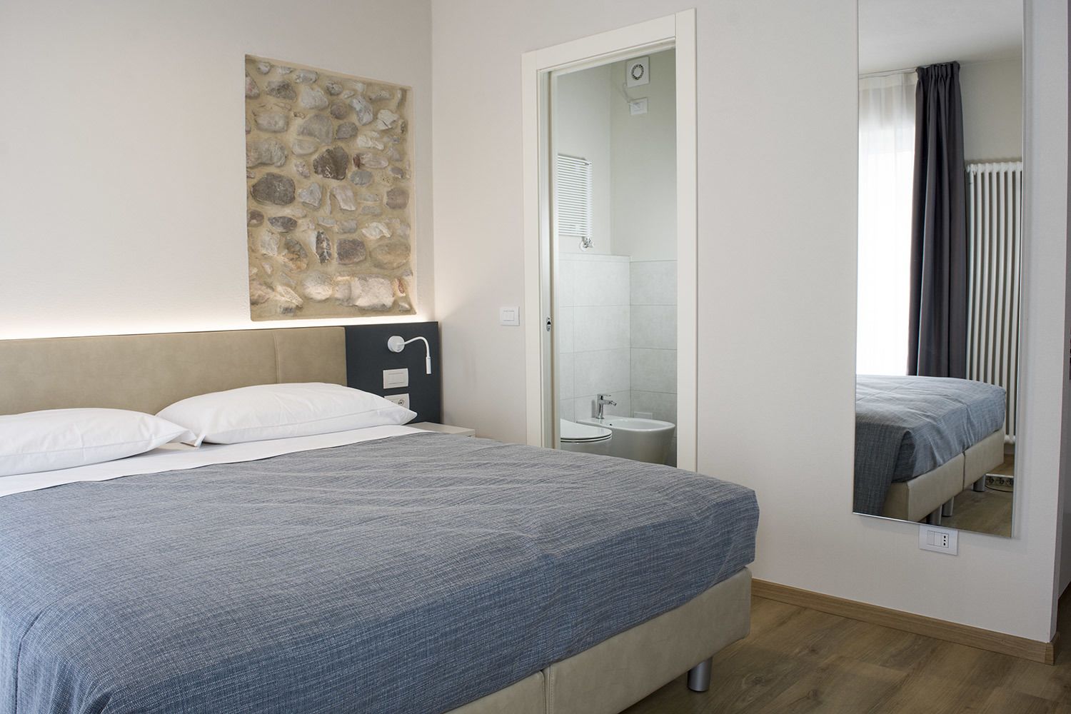 A bedroom with a bed, a nightstand, a mirror, and a stone wall.