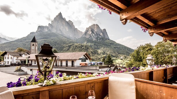 A restaurant with a wooden deck overlooking a mountainous landscape, featuring a bell tower and a view of the mountains. - Enzian Genziana (Castelrotto)