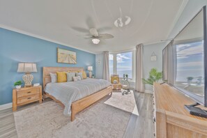 Ein Schlafzimmer mit Bett, Nachttischen, einer Kommode, einem Deckenventilator, einem großen Fenster mit Vorhängen und Blick zum Himmel.