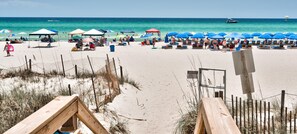 A beach with multiple umbrellas and beach chairs, a wooden fence, and a sign. - Emerald Beach Penthouse #2525 2BR+3 Bunks 2 BA Remodeled Summer Deals (Panama City Beach)