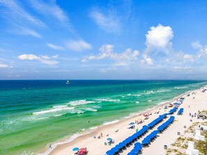 Ein Strand mit klarem türkisblauem Wasser, einer Pier in der Ferne und Reihen blauer Sonnenschirme am Ufer.