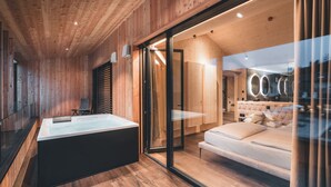A modern room with wooden paneling, a large bathtub, a sofa, and a glass wall offering a view of the bathroom.
