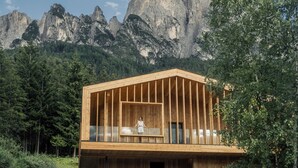 A wooden cabin with a balcony, set against a mountainous backdrop.
