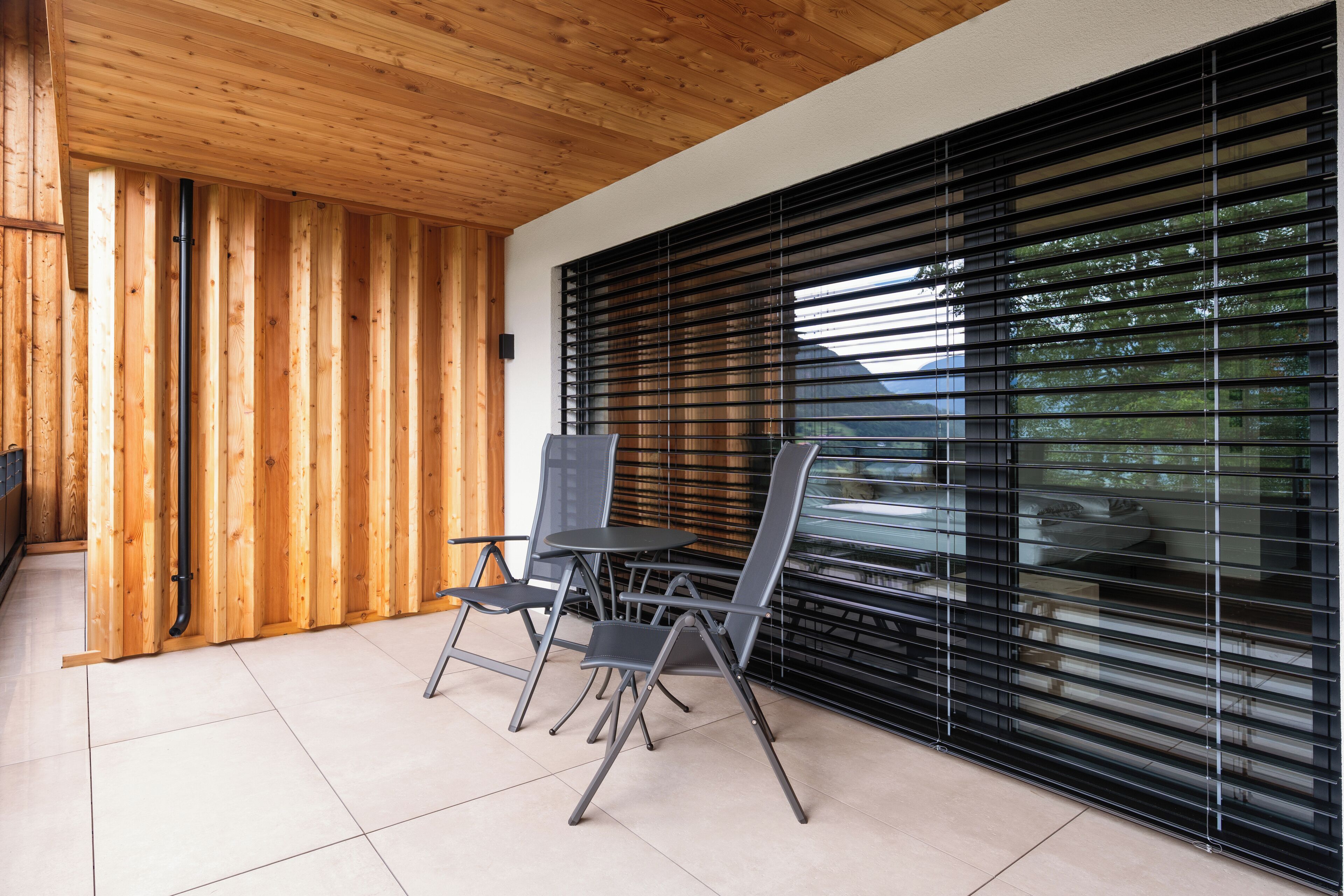 A modern outdoor seating area with a wooden wall and black blinds, featuring a table and chairs.